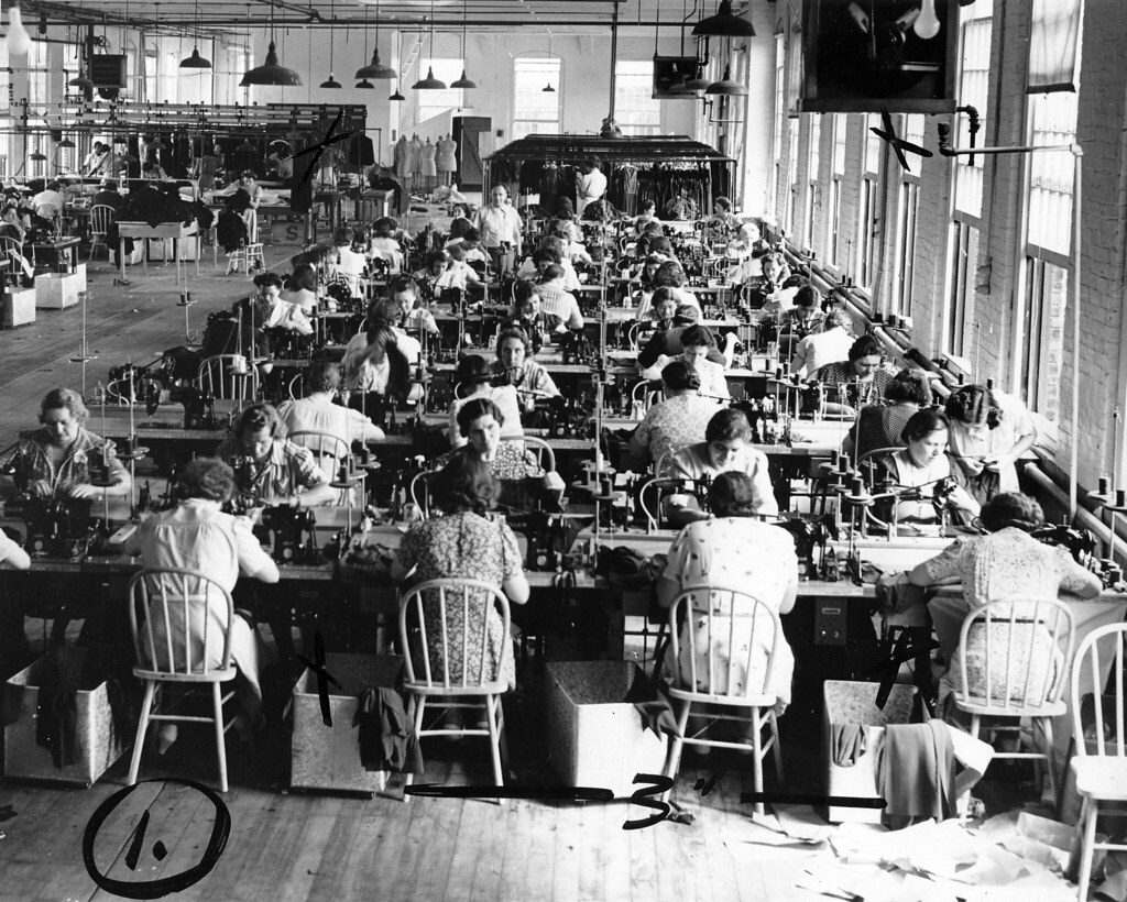 Women sewing at long tables beside tall windows in a garment factory